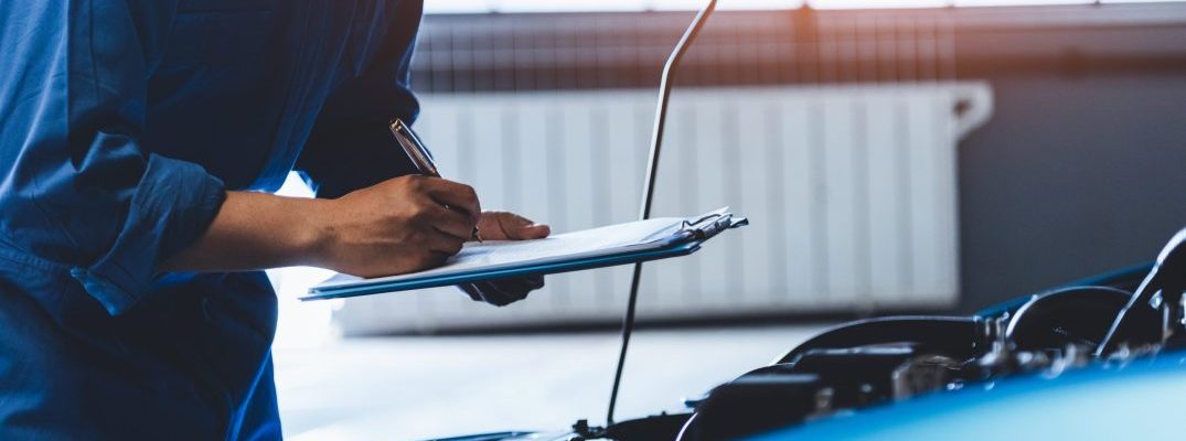 A technician with a clipboard inspects under the hood of a car.